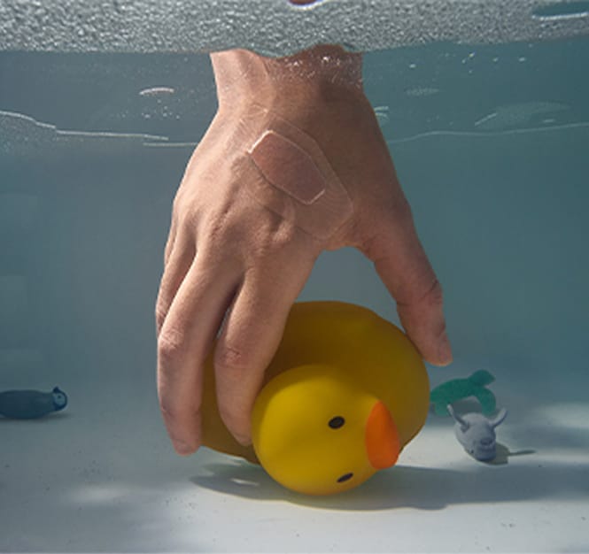 Uma mão com um curativo, submersa na água, segurando um grande pato de borracha amarelo, cercada por miniaturas de animais de brinquedo flutuando.
